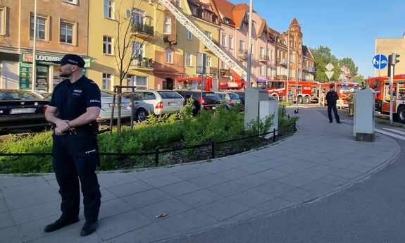 na pierwszym planie policjant stojący na chodniku, w tle wozy strażackie przy budynku